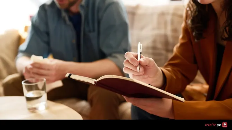 A woman writes in a notebook during a conversation with a man sitting on a sofa beside a wooden table with a glass of water
